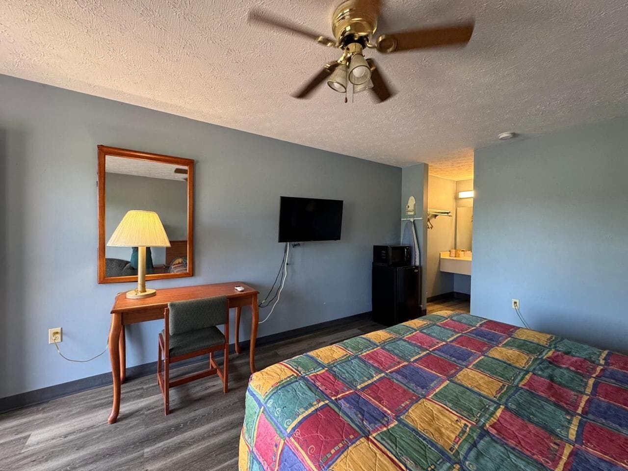 Habitación de hotel con cama, escritorio, silla, ventilador de techo, televisor y espejo.