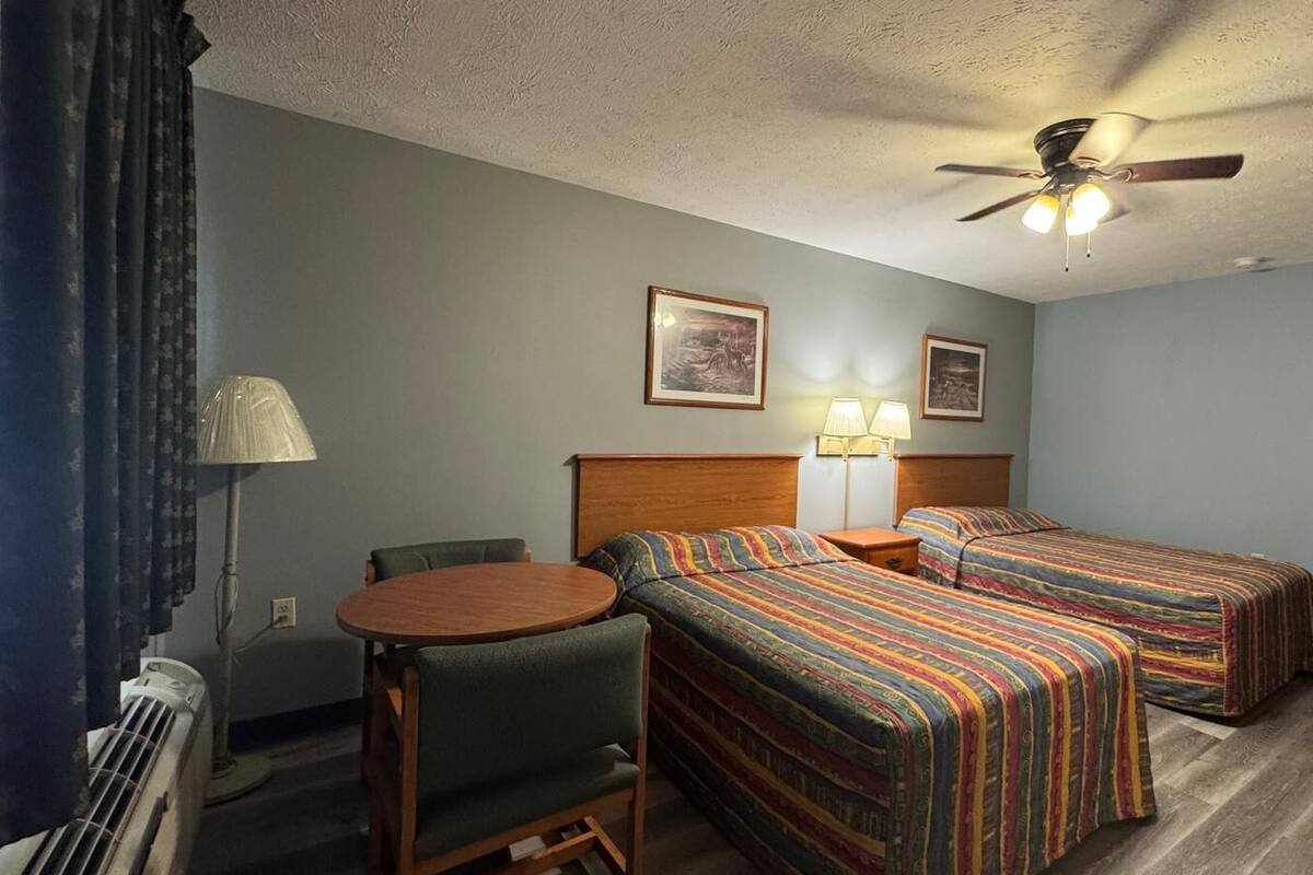 Habitación de hotel con dos camas, una mesa redonda, una silla, un ventilador de techo y un espejo.