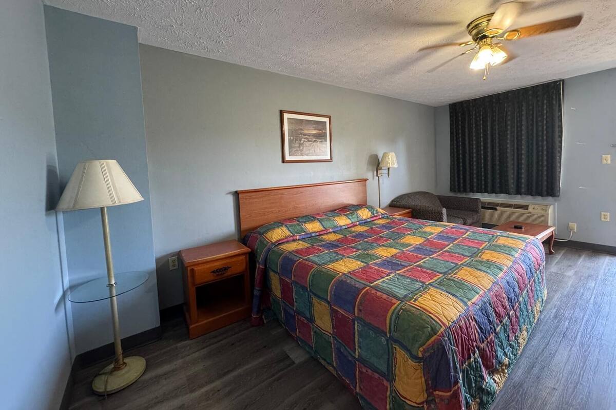 Habitación de hotel con una cama de madera, un ventilador de techo, una mesita de noche, una lámpara, un cuadro enmarcado en la pared y una ventana con cortinas.