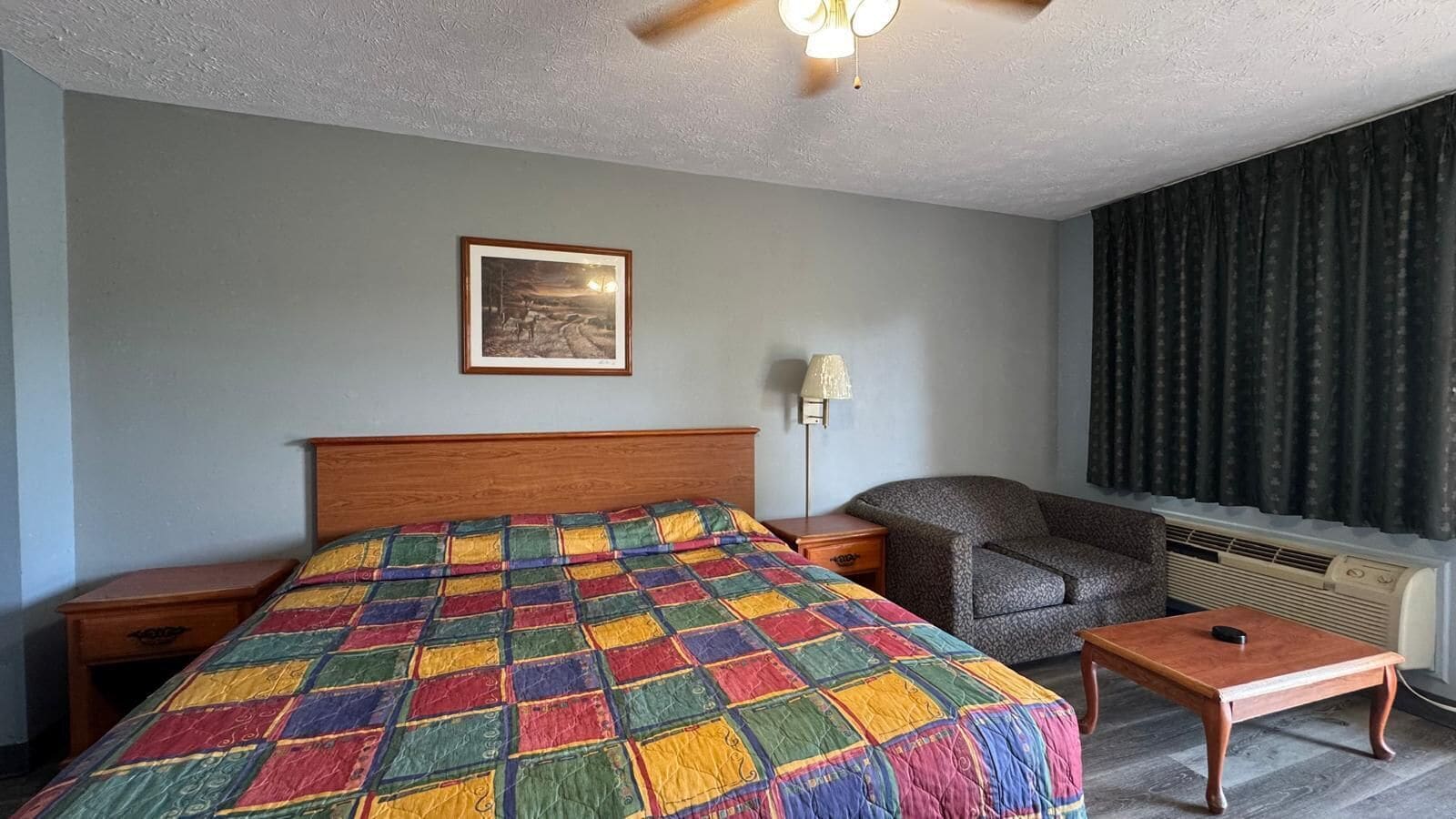 A hotel room with a bed, a nightstand, a chair, a table, a lamp, a picture on the wall, and a window with curtains.