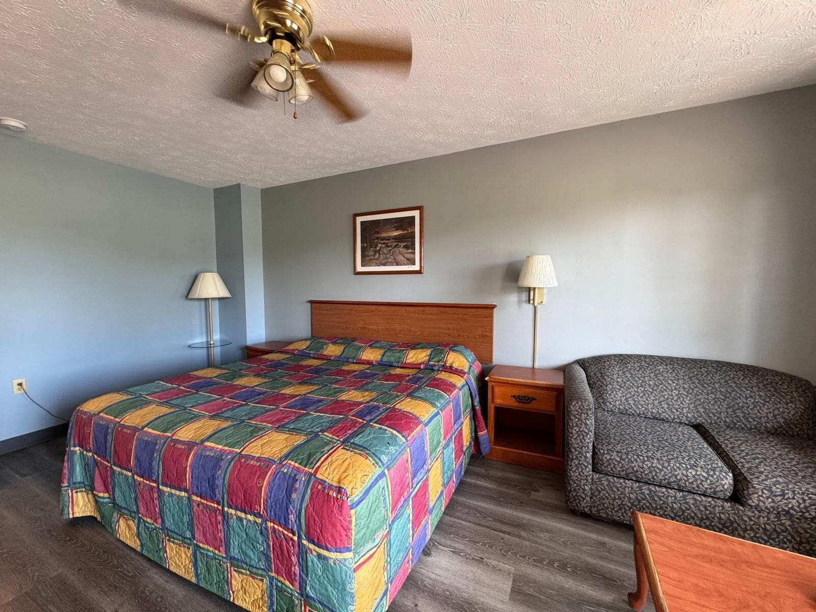 A hotel room with a wooden bed, a gray armchair, a small wooden table, a ceiling fan, and a framed picture on the wall.
