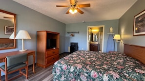 A hotel room with a bed, a desk with a lamp, a television, a wardrobe, and a ceiling fan.