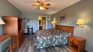 A hotel room with a bed, a desk, a chair, a ceiling fan, a trash can, and a nightstand with a lamp.