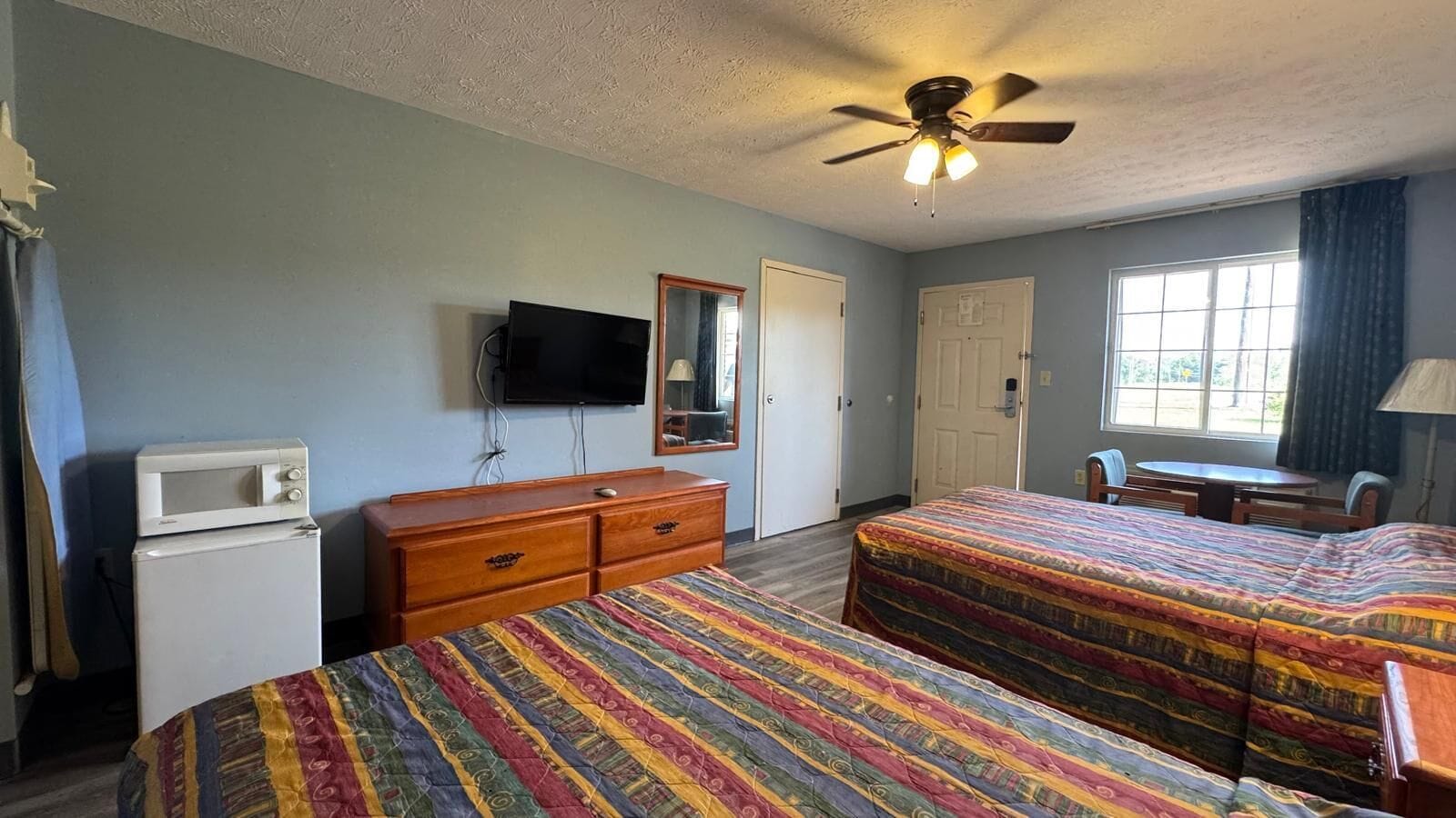 A room with two beds, a television, a microwave, a ceiling fan, and a window with curtains.