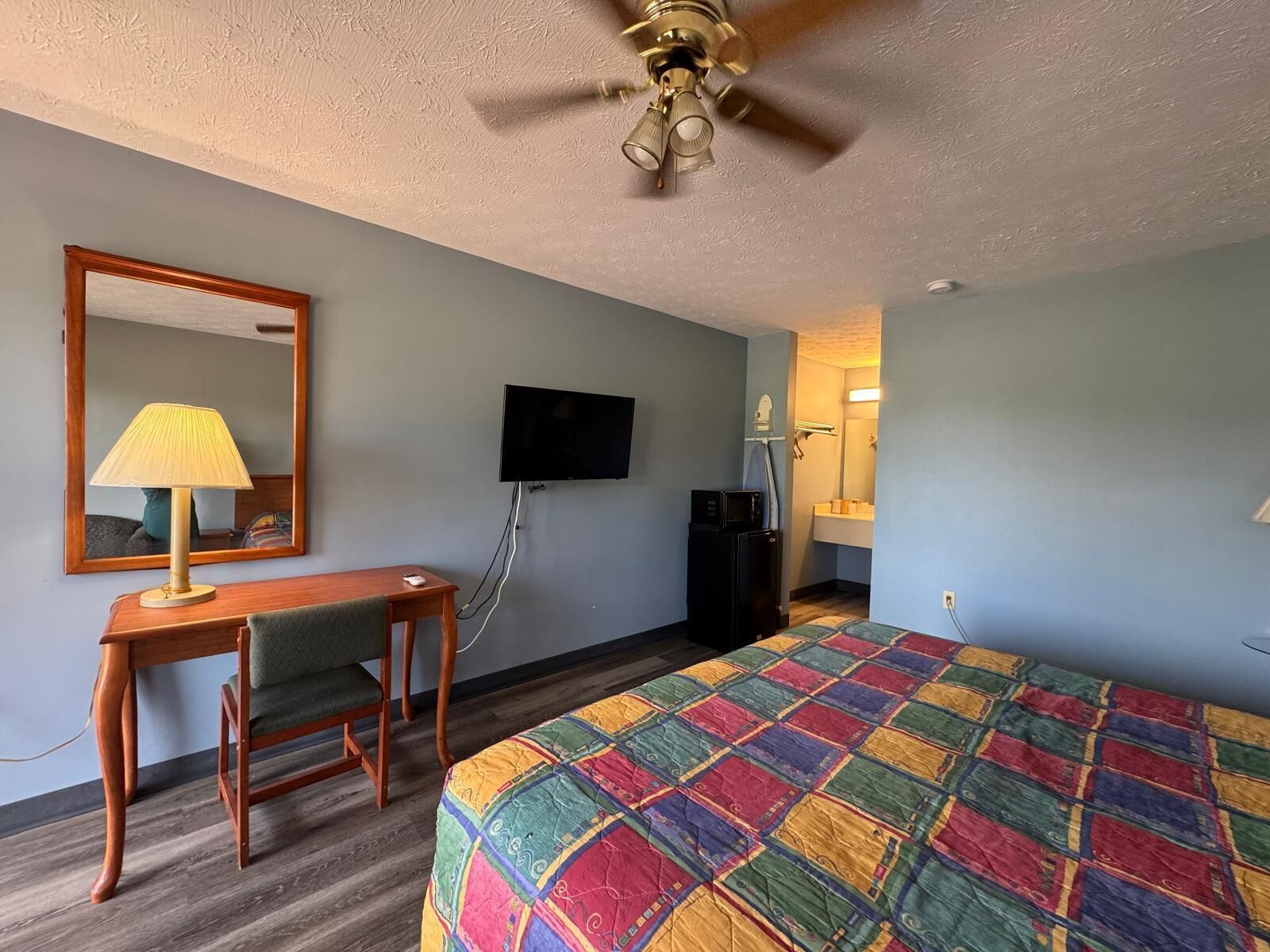A hotel room with a bed, a desk, a chair, a television, a ceiling fan, and a bathroom visible through an open door.