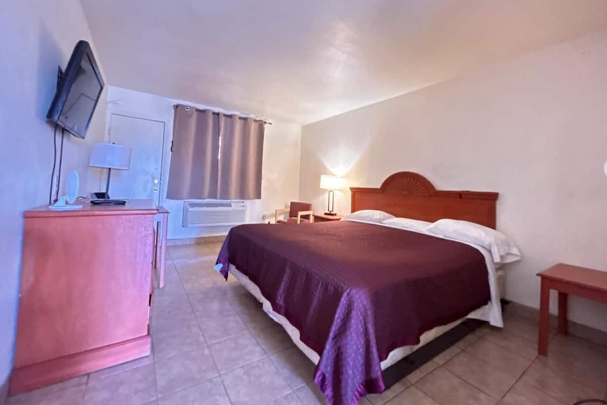 A hotel room with a bed, bedside table, lamp, TV, and a desk with a chair.