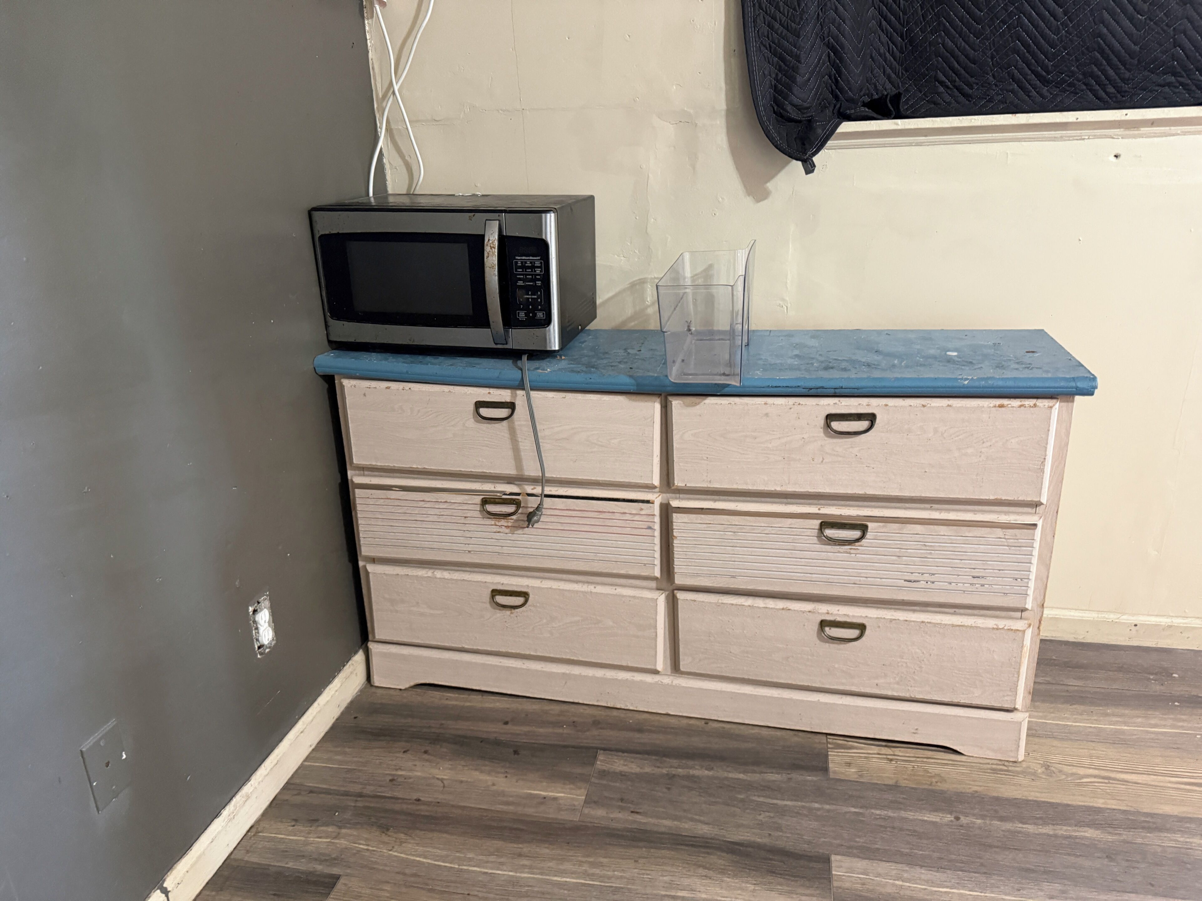 Una kitchenette con un microondas sobre una encimera azul, un aparador blanco y un recipiente de plástico transparente.