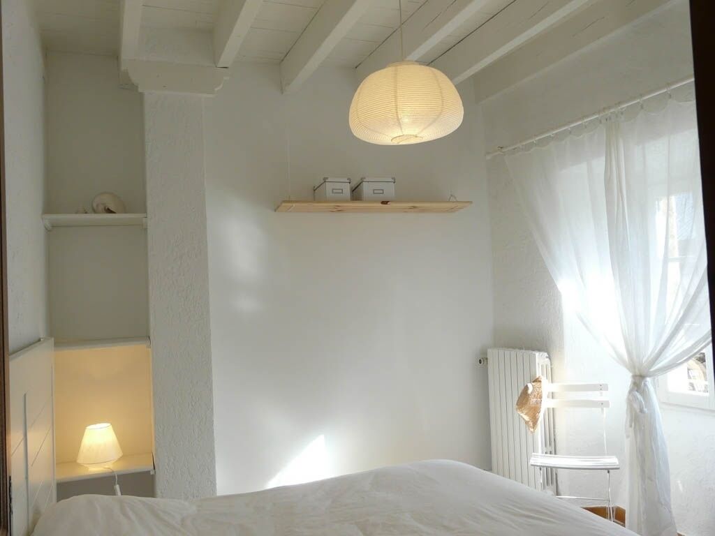 A bedroom with a bed, a hanging light fixture, a shelf with storage boxes, a radiator, and a window with curtains.