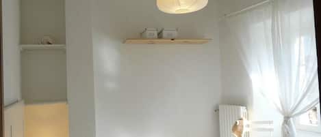 A bedroom with a bed, a hanging light fixture, a shelf with storage boxes, a radiator, and a window with curtains.