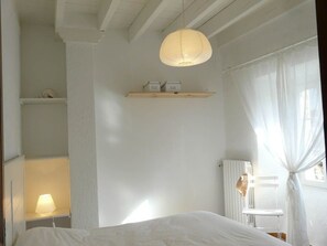 A bedroom with a bed, a hanging light fixture, a shelf with storage boxes, a radiator, and a window with curtains. - Sainte Germaine Vacation Rental "La Ferme (8 people)" (LUC)