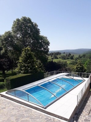 Una piscina con cerramiento de vidrio, rodeada de pavimento de piedra, con árboles y colinas como telón de fondo.