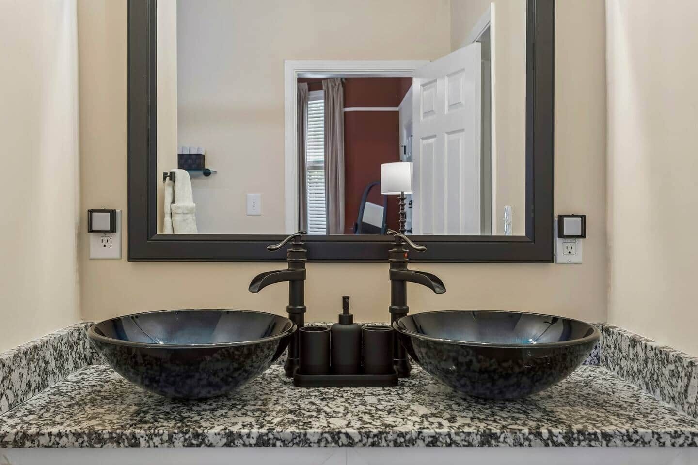 A bathroom with two large bowls on a granite countertop, a mirror above, and a door visible in the reflection.