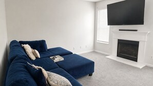 Ein Wohnzimmer mit einem blauen Ecksofa, einem flachbildfernseher über einem Kamin und einem Fenster mit Jalousien.