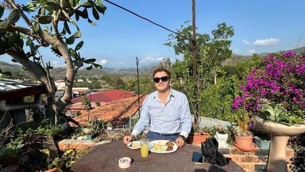Una persona sentada en una mesa al aire libre con un plato de comida y una bebida, rodeada de plantas en macetas y con vistas a un edificio de tejados rojos.