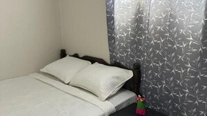 Una cama individual con cabecera de madera, un almohadón blanco y una mesita de noche con una botella y una taza.