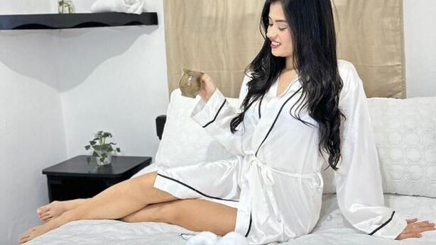 A person sitting on a bed in a room with a black nightstand, a white pillow, and a beige curtain. - Guest House Don Moisés (Copan Ruinas)