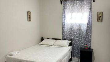 A single bed with a white bedspread and pillows, a small bedside table with a phone, and a window with a patterned curtain.