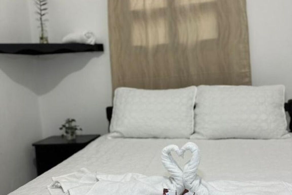 Una cama bien hecha con ropa blanca, una disposición de toallas en forma de corazón, una mesita de noche con una planta y un cabecero de madera.