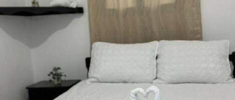 Una cama bien hecha con ropa blanca, una disposición de toallas en forma de corazón, una mesita de noche con una planta y un cabecero de madera.