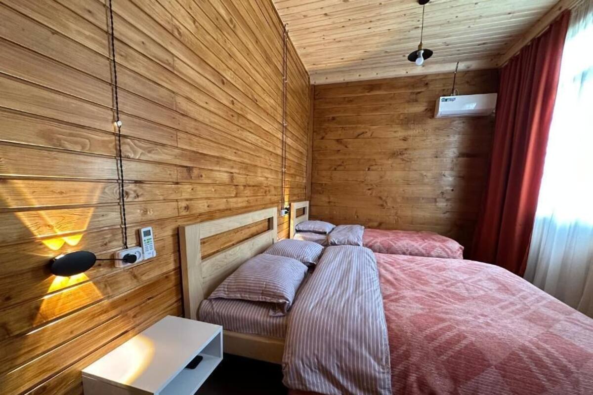 A wooden-paneled room with two beds, a bedside table, a wall-mounted heater, and a window with curtains.