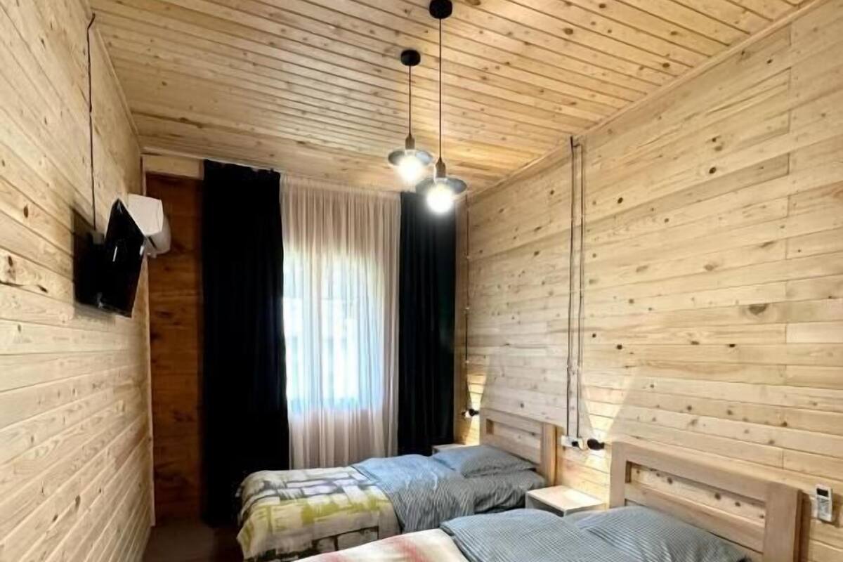 A wooden-paneled room with two beds, a ceiling light, and a window with curtains.
