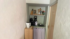 Eine kleine Küche mit Mikrowelle, Kaffeemaschine und Geschirr auf einem Regal. Im Eck befindet sich ein Holzschrank und eine Bank.