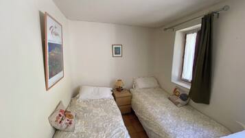 A small, single-room accommodation with two beds, a nightstand, a lamp, a window with curtains, and a framed picture on the wall.