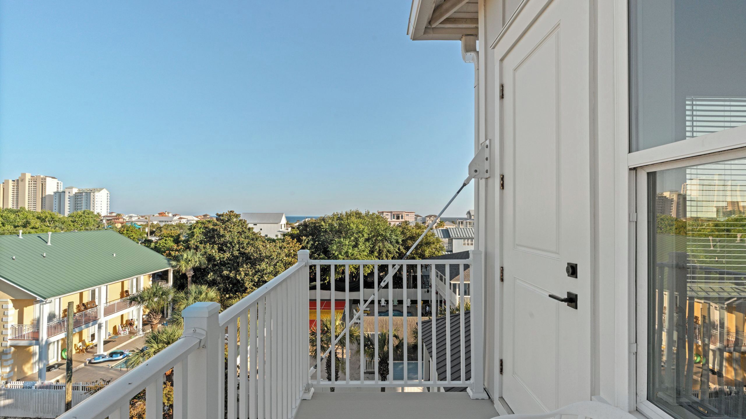 A balcony with a white railing overlooking a residential area with trees and a building with a green roof.
