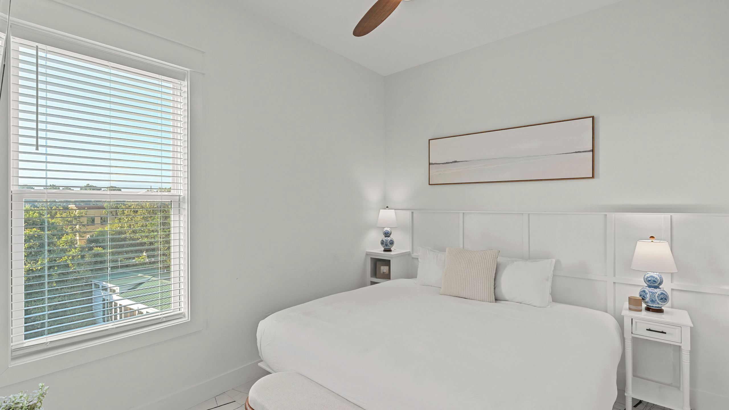 A bedroom with a large bed, two bedside tables with lamps, a ceiling fan, and a window with blinds.