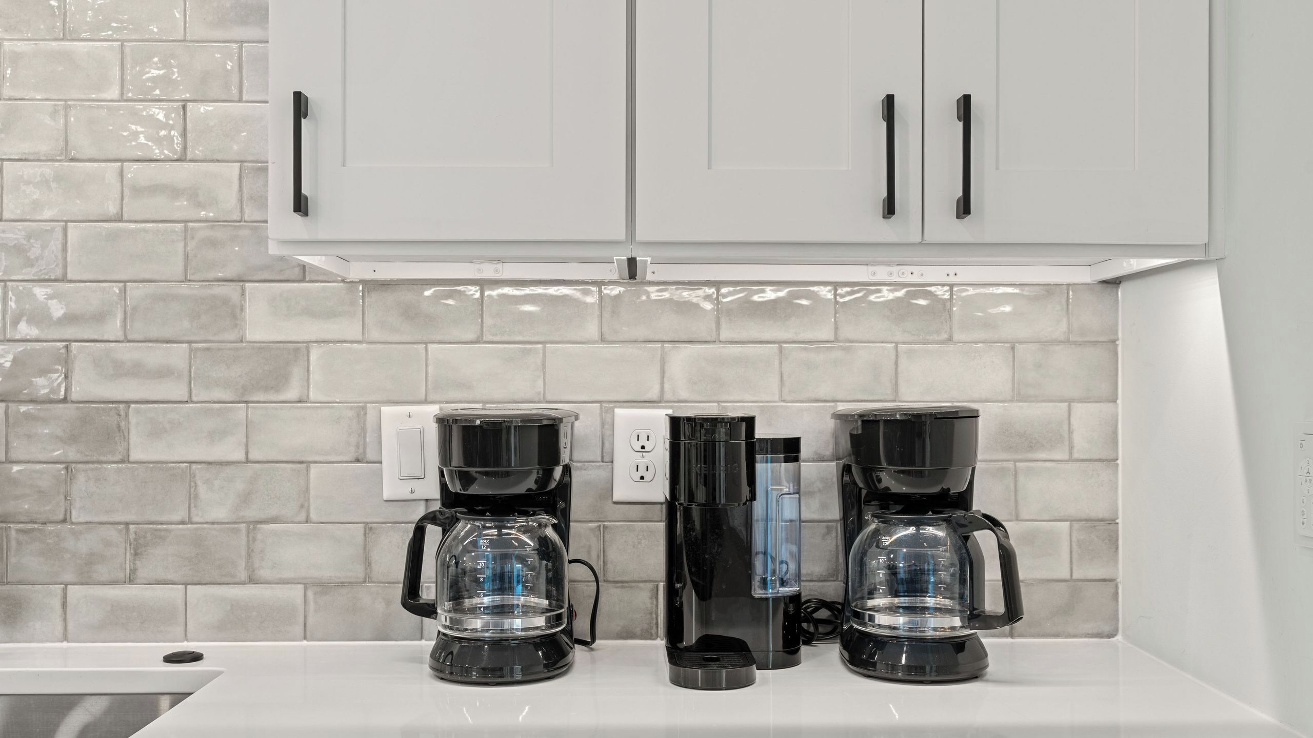 A kitchen with white cabinets, a white countertop, and two coffee makers.