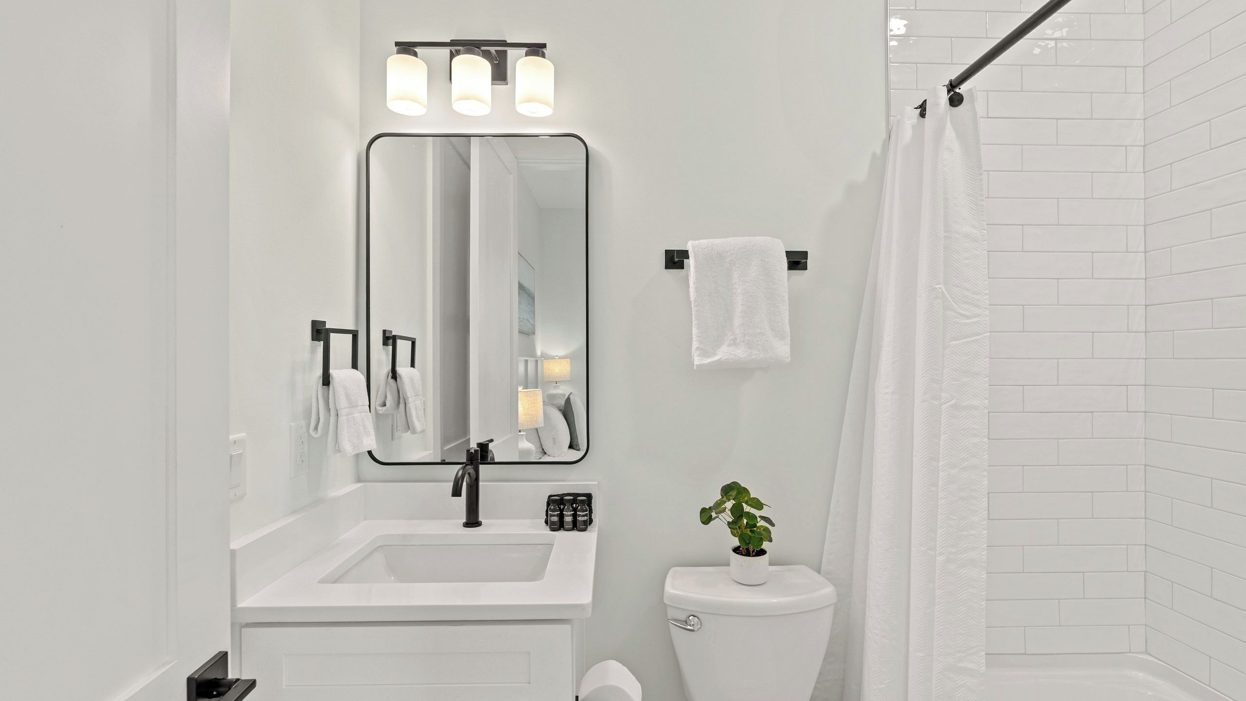 A modern bathroom with a white sink, black faucet, and a large mirror above it. There is a toilet with a plant on top and a shower curtain.
