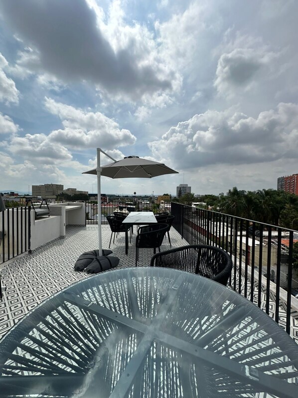 Una terraza en la azotea con una mesa de vidrio, sillas negras y un paraguas blanco.