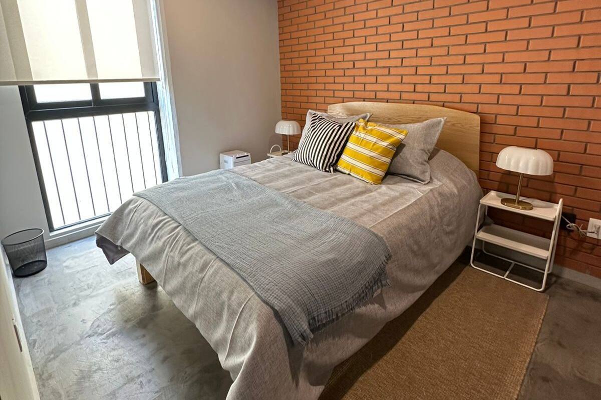 Un dormitorio con pared de ladrillo, una cama con colcha gris, una ventana con persianas y una mesita de noche con lámpara.