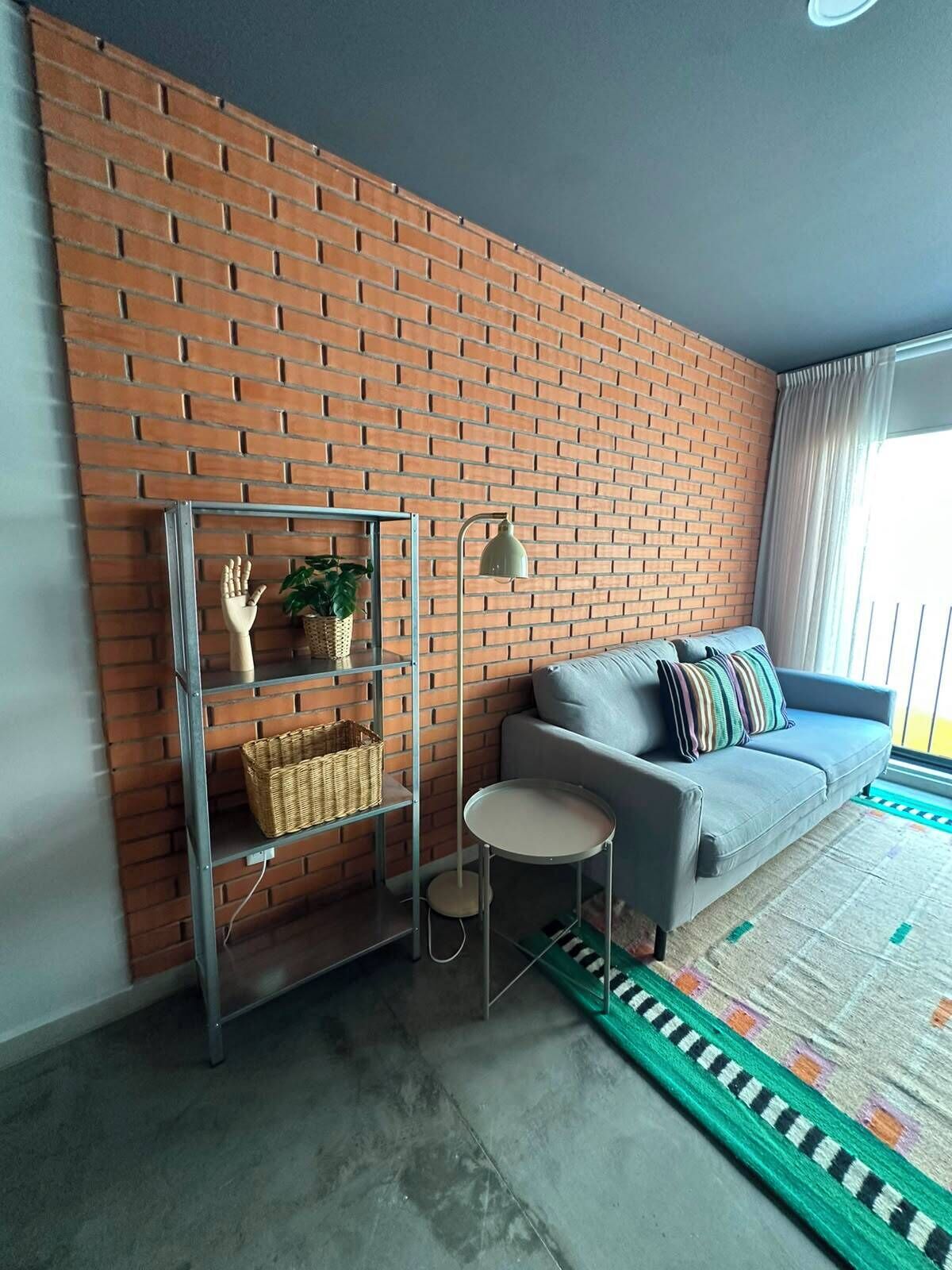 A living room with a brick wall, a grey sofa with patterned cushions, a small round table, and a metal shelving unit with baskets.