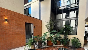 Un patio con piso de ladrillo, una banca, plantas en macetas y un edificio con grandes ventanales y balcones.