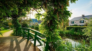 Un pont vert enjambe un étang, et un sentier mène à un bâtiment à la façade blanche et aux volets verts.