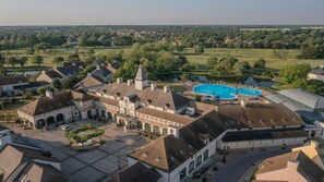 Luftaufnahme einer Wohngegend mit großem Schwimmbecken, mehreren Gebäuden unterschiedlicher Architektur und umgebender Bepflanzung.