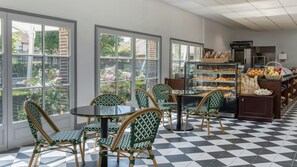 Un café avec de grandes fenêtres, des chaises en osier, un sol carrelé et une vitrine remplie de pâtisseries.