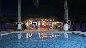 Une piscine dont le fond est recouvert de carreaux formant un motif géométrique, entourée d’une clôture, de palmiers et d’un bâtiment doté d’une porte rouge et de lumières.