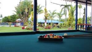 Una mesa de billar con un repuesto de bolas, una vara y vista a un jardín con palmeras y un edificio al fondo.