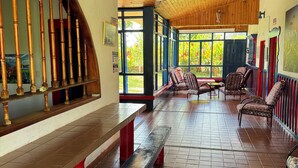 Un espacio de estar al aire libre cubierto con bancos y sillas de madera, suelo de baldosas y amplias ventanas que dejan entrar luz natural.