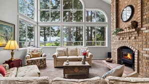 A cozy living room with a fireplace, large windows, and comfortable seating. - Cedar & Sound by Avantstay Indoor Hot Tub + Game Room + Media Room Sound + Olympic Mtn Views (Freeland)