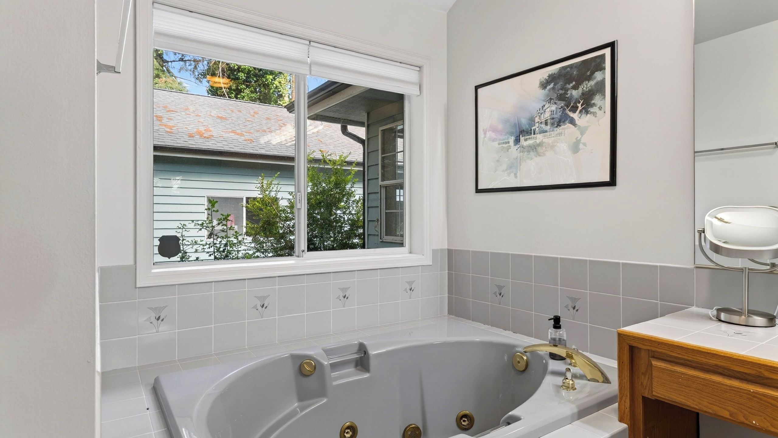 A bathroom with a large bathtub, a window with a view of a house, and a framed picture on the wall.