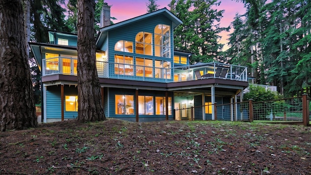 A two-story house with large windows, a balcony, and a forested backdrop. - Cedar & Sound by Avantstay Indoor Hot Tub + Game Room + Media Room Sound + Olympic Mtn Views (Freeland)