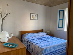 A bedroom with a wooden bed, a small bedside table with a lamp, a wall-mounted picture, and a window with a blue frame. - A typical fisherman's cottage. (Favignana)