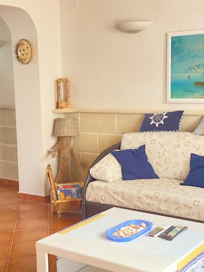 A cozy living room with a sofa, a small table with a bowl of seashells, a lamp, and a framed picture on the wall. - A typical fisherman's cottage. (Favignana)