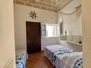 A room with two beds, a wooden door, a window with blue shutters, a wall-mounted shelf, and a decorative wall piece. - A typical fisherman's cottage. (Favignana)