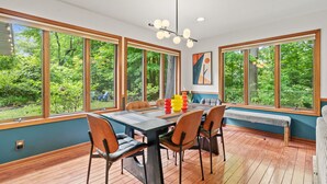 A dining area with a table, chairs, and a bench, large windows with a view of greenery, and a ceiling light fixture. - Secluded Mid-Century Haven | Hot Tub | Fire Pit (Fennville)