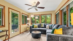 A spacious room with a large gray sectional sofa, a round ottoman, a ceiling fan, and large windows offering a view of greenery. - Secluded Mid-Century Haven | Hot Tub | Fire Pit (Fennville)