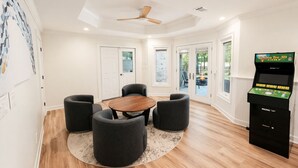 Um quarto com uma mesa redonda de madeira, quatro poltronas cinzentas, um fliperama preto e um ventilador de teto.